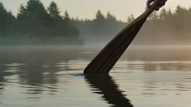 Ein Holzpaddel taucht vor bewaldetem Ufer ins ruhige Wasser