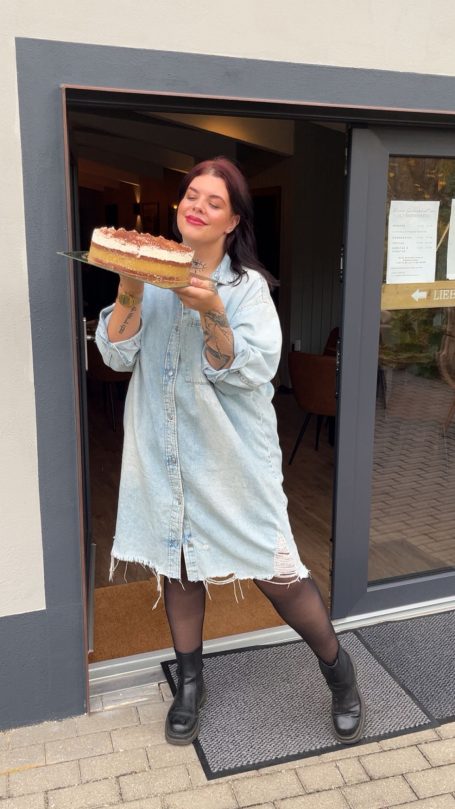 Frau in Jeansjacke steht vor einer Tür und hält eine Torte in den Händen.