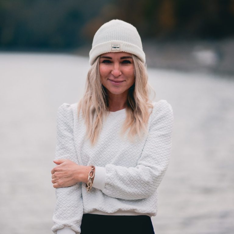 Junge Frau mit blonden Haaren und Mütze steht am Wasser und lächelt.