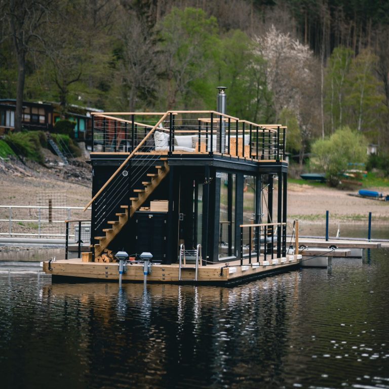 Modernes schwimmendes Haus mit Holzsteg und Treppe, umgeben von Wasser und Bäumen.