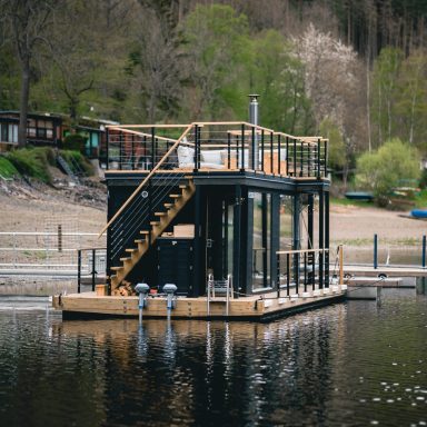 Schwarzes Haus auf dem Wasser mit Treppe und umlaufendem Balkon, umgeben von Bäumen.