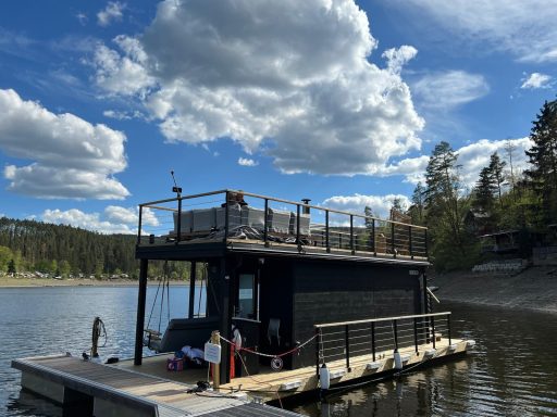 Schwarzes schwimmendes Haus auf ruhigem Wasser, umgeben von Bäumen und Wolken.